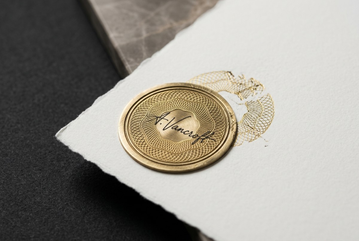 An extreme macro detail of a single document corner on charcoal: a raised brass register seal half-overlapping a signature, where delicate g