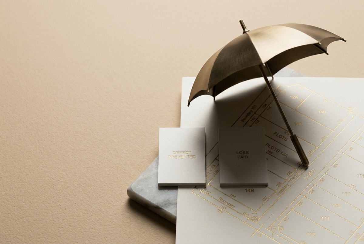 A macro overhead flatlay on warm sand-toned paper of a delicate brass umbrella, opened and tilted, casting its curve over a finely engraved